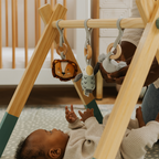 A baby lies on a play mat reaching up toward the Wooden Play Gym with hanging toys, while an adult adjusts one of the toys. A crib can be seen in the background.