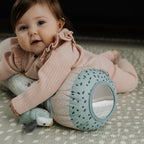 A baby in a pink outfit lies on a patterned mat, hugging the Animal Adventures Tummy Time Pillow—a sensory baby pillow with a mirror and animal design—while looking up with a gentle expression.