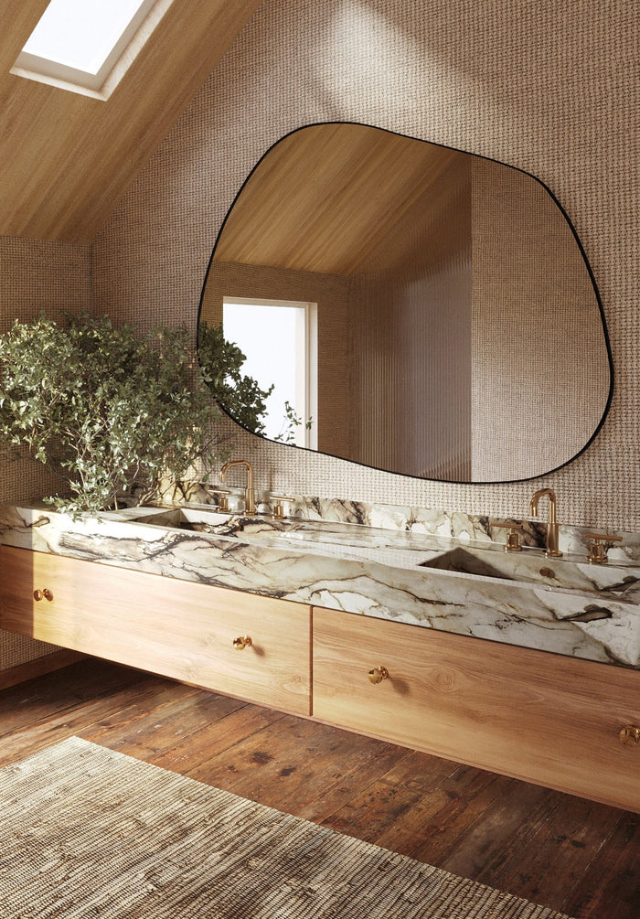 Asymmetrical wall mirror mounted above a double marble bathroom vanity with brass fixtures and wood cabinetry in a warm, naturally lit space.