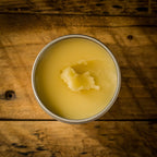 Top view of an open tin of Mammoth Beard Co. beard balm on a rustic wooden surface, highlighting its smooth, natural formula with a small portion scooped out.