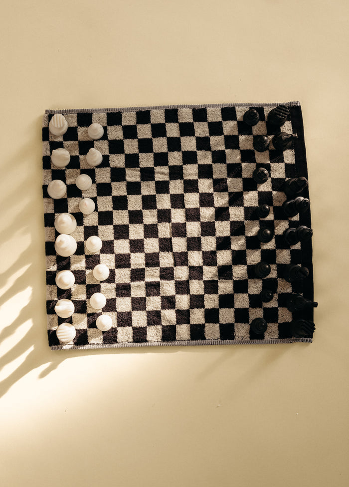 Black and white checkered towel laid flat on a surface, styled as a chessboard with white and black chess pieces arranged on top to highlight the geometric pattern and texture.
