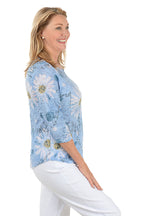 Woman wearing a blue floral blouse with white pants on a white background. Side shot.