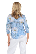 Woman wearing a blue floral blouse with white pants on a white background. Back shot.