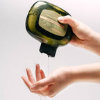 Hand using a soap dispenser to apply liquid soap to another hand against a light background.