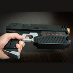 Hand holding a black and silver Judge Dredd Lawgiver cosplay prop gun with orange and red accents on a dark wood surface.