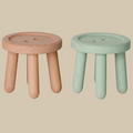 Two small round wooden stools with three legs, shown side by side on a plain light beige background. The left stool is unfinished natural wood, and the right stool has a distressed sage green paint finish.