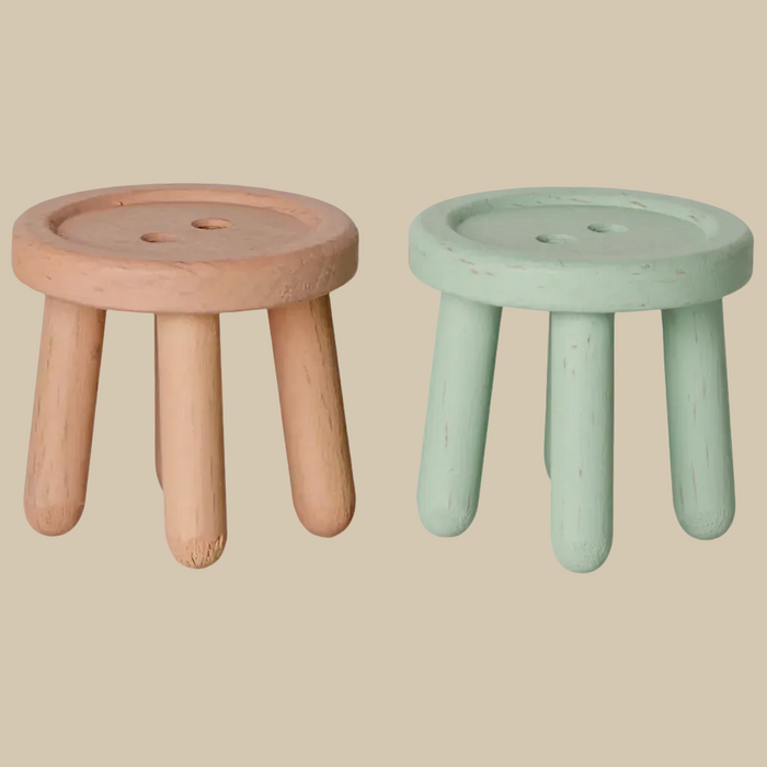 Two small round wooden stools with three legs, shown side by side on a plain light beige background. The left stool is unfinished natural wood, and the right stool has a distressed sage green paint finish.
