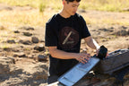 A person wearing a black Leatherman Blade & Grain tee pulling a camera out of a camera pack.