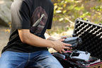 A person pulling a drone out of a carrying case while wearing a black Leatherman Blade & Grain Tee with a fixed blade design on the front.