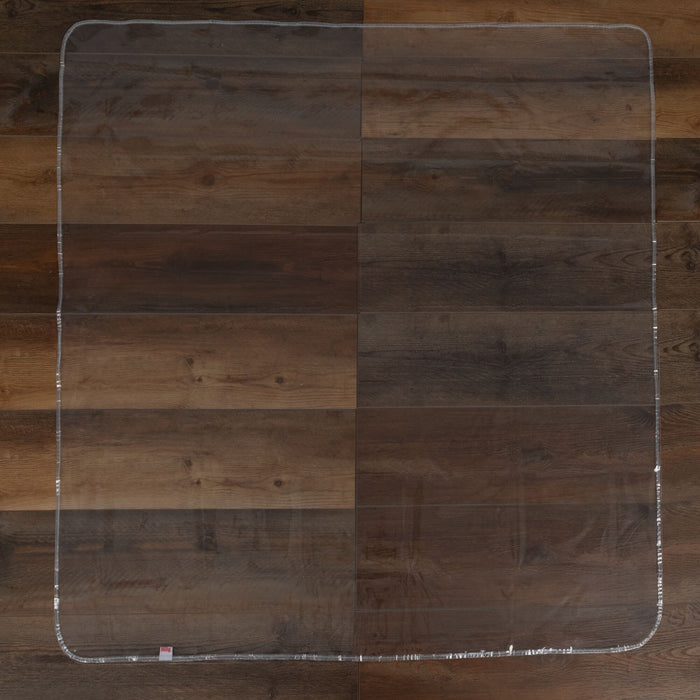 The Nuby US No-Mess Mealtime Mat, a clear rectangular floor protector with rounded corners, is laid flat on dark brown hardwood flooring; easy to clean for effortless maintenance.