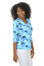 Woman wearing a blue palm tree patterned blouse on a white background. Side shot.