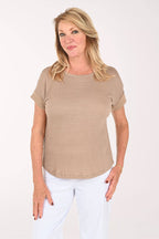 A woman modeling a short-sleeve, crew-neck knit top in a light taupe or beige color, paired with white pants, against a plain white studio background.