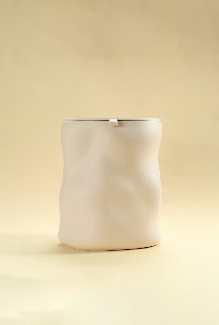 A beige sculptural dustbin with a fitted lid, photographed against a light yellow background.