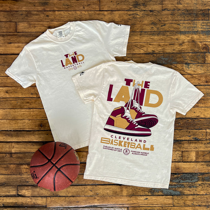 Two white t-shirts with 'The Land' and basketball graphics on a wooden surface, accompanied by a basketball.