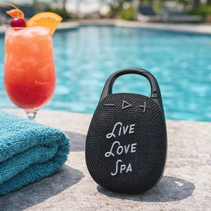 Wireless Clip Speaker | Live Love Spa in black with textured surface, placed by a pool next to a blue towel and a tropical drink, showcasing a portable wellness device for outdoor use.